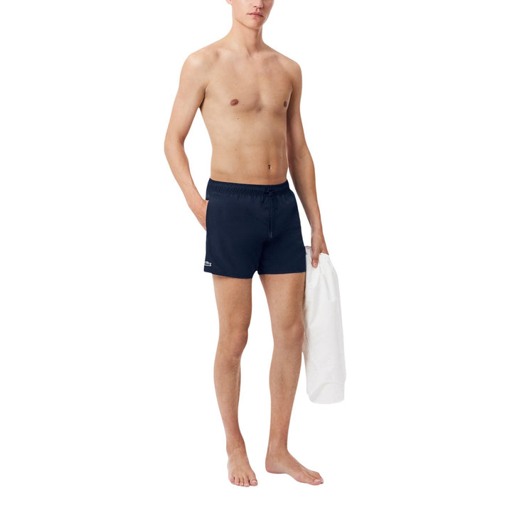 Image 1 : Homme portant un short de bain Lacoste bleu marine coupe courte avec logo crocodile brodé, vue de face, tenant une serviette blanche — Delvale