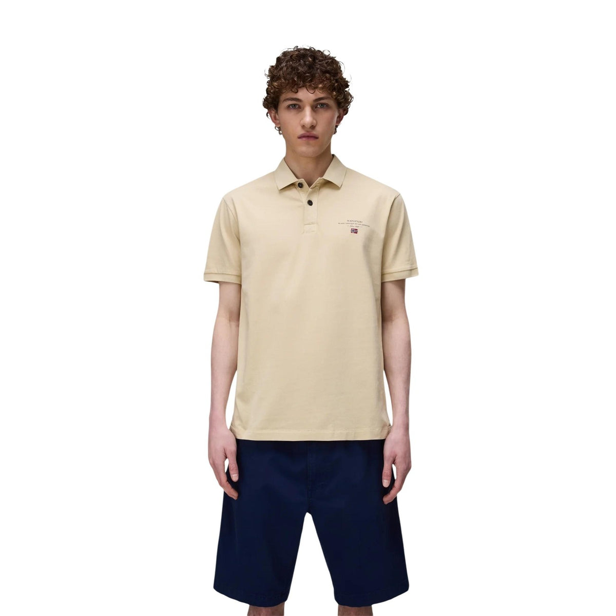 Image 1 : Homme portant le polo Napapijri manches courtes en coton piqué coloris sable, avec logo brodé et drapeau norvégien côté poitrine, associé à un bermuda bleu marine — Delvale