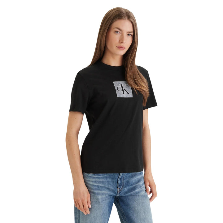 Image 1 : Femme portant le t-shirt noir Calvin Klein Jeans avec logo CK carré argenté sur la poitrine, associé à un jean bleu, vue de trois quarts — Delvale