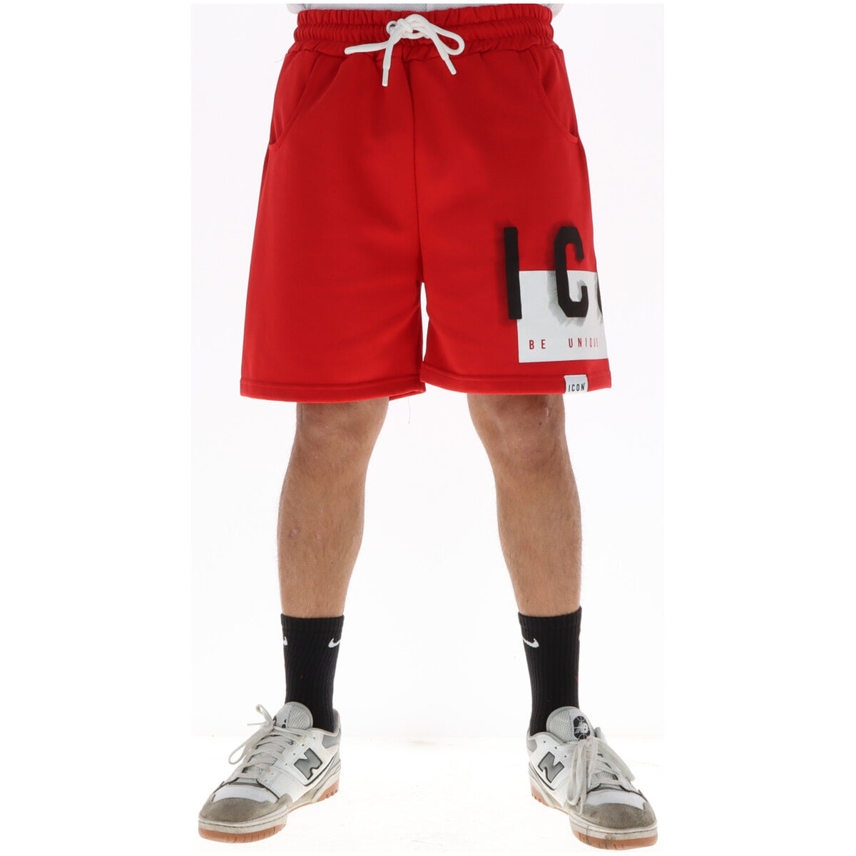 Image 1 : Bermuda homme Icon rouge avec logo oversize Be Unique noir et blanc, taille élastique avec lacet blanc, porté avec baskets New Balance grises — Delvale