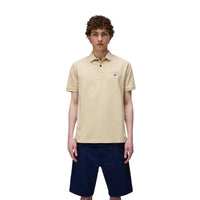 Image 1 : Homme portant le polo Napapijri manches courtes en coton piqué coloris sable, avec logo brodé et drapeau norvégien côté poitrine, associé à un bermuda bleu marine — Delvale